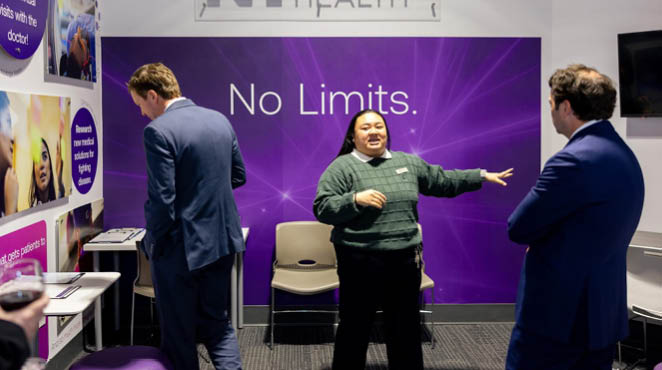 A woman in a green shirt stands in front of a purple sign that says “No Limits." Two men are standing behind her, one on the left and one on the right. They are all in a room with a dining table and chairs. AI generated content