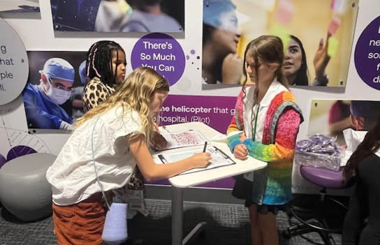 Three girls are standing around a table, one of them signing a book. The girls are wearing name tags. AI generated content