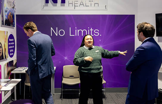 A woman in a green shirt stands in front of a purple sign that says “No Limits." Two men are standing behind her, one on the left and one on the right. They are all in a room with a dining table and chairs. AI generated content