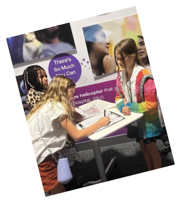 Three girls are standing around a table, one of them is signing a book. AI generated content